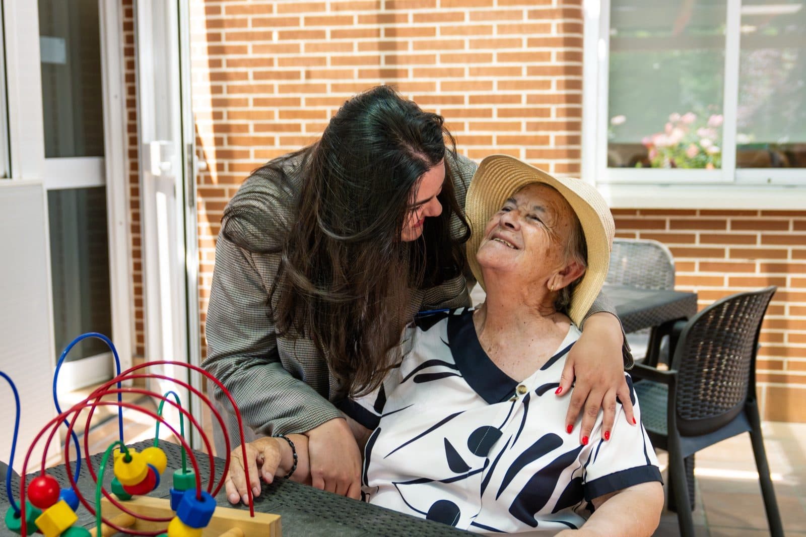 Cuidadora abrazando a una señora mayor con sombrero en la terraza de su casa